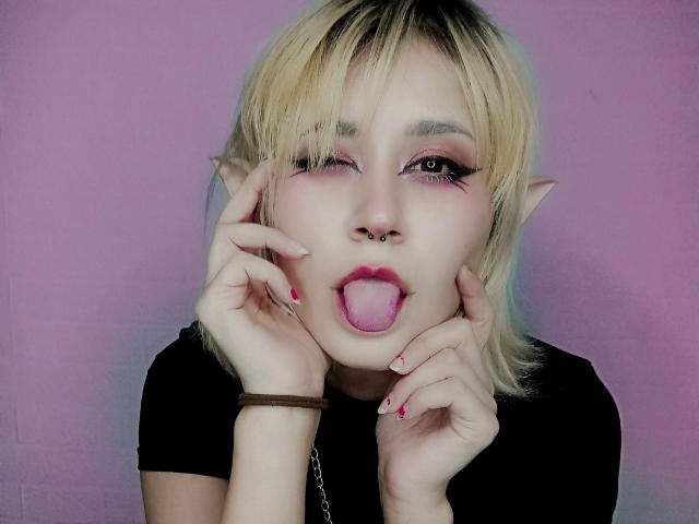 AlexaLexie mit blonden Haaren, spitzen Elfenohren, Nasenring, pinkem Eyeliner und herausgesteckter Zunge, die ihr Gesicht mit lackierten Nägeln berührt, vor einem lila Hintergrund..