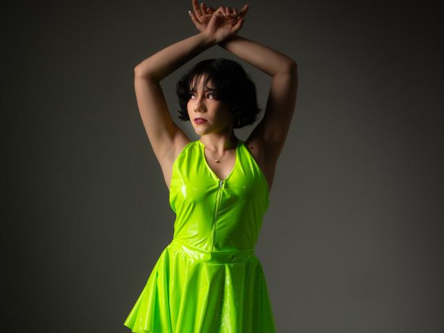 Sharonnyx con le braccia alzate, indossa un abito in lattice verde neon lucido, capelli neri corti, guarda leggermente di lato, sfondo grigio, illuminazione soffusa, cerniera dell'abito leggermente aperta..