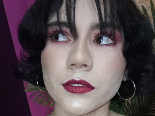 Sharonnyx hat eine blasse Haut, dunkles, lockiges Haar, roten Eyeliner und roten Lippenstift. Sie trägt Ohrringe im Hula-Hoop-Stil mit einer leichten Aufwärtsbiegung und blickt zur Seite mit einem neutralen Gesichtsausdruck..