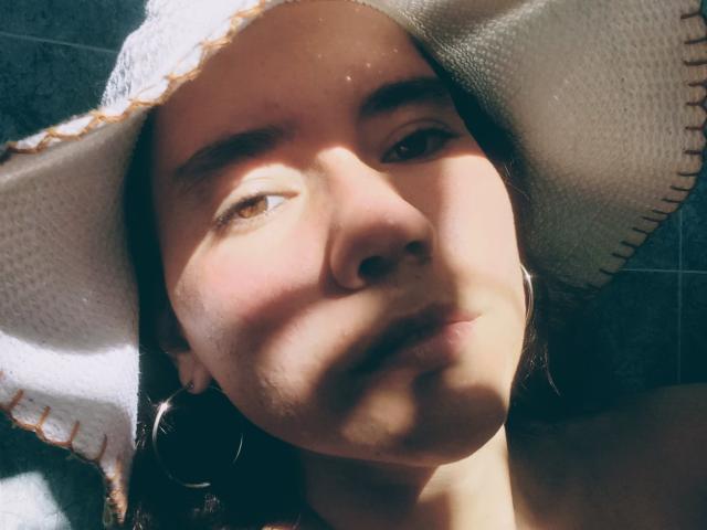 O rosto de Sharonnyx com um chapéu de sol, sombras na pele, olhos castanhos olhando ligeiramente para cima, nariz pequeno, lábios levemente entreabertos, cabelo escuro, brincos de argola, luz suave, expressão casual..