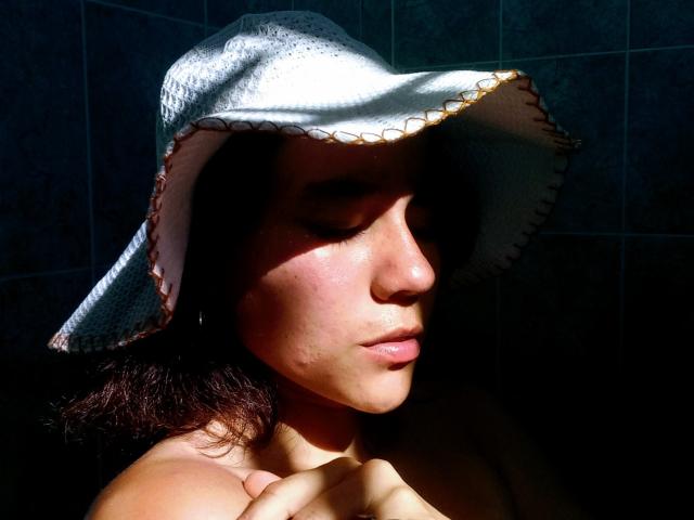 Sharonnyx, com o rosto iluminado pelo sol, usando um chapéu branco com detalhes dourados, tem a pele úmida e cabelos escuros, segurando o ombro nu, em um fundo sombrio com azulejos escuros..