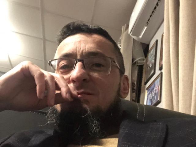BernardFunny has a bearded face with glasses, dark hair, and a pensive expression, resting his chin on his hand, wearing a dark plaid shirt, in a selfie.
