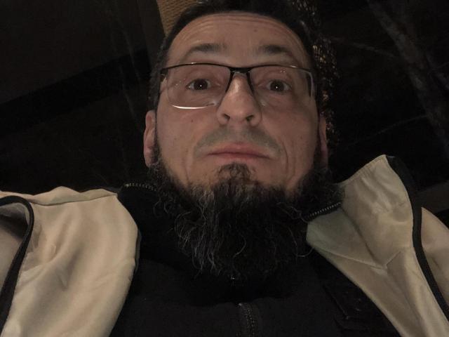 BernardFunny, with glasses, a beard, and a serious face, wearing a beige jacket over a black vest, looking into the camera. His nose is slightly upturned.