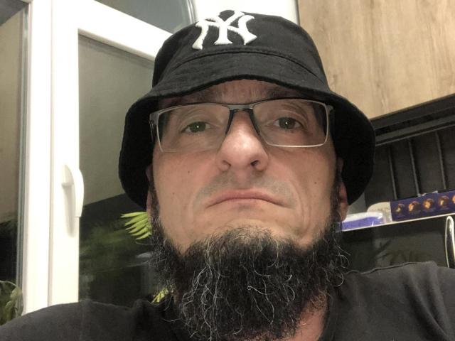 BernardFunny with a black baseball cap, white New York Yankees logo, glasses, and a thick, curly beard, looking serious. He's wearing a black shirt.