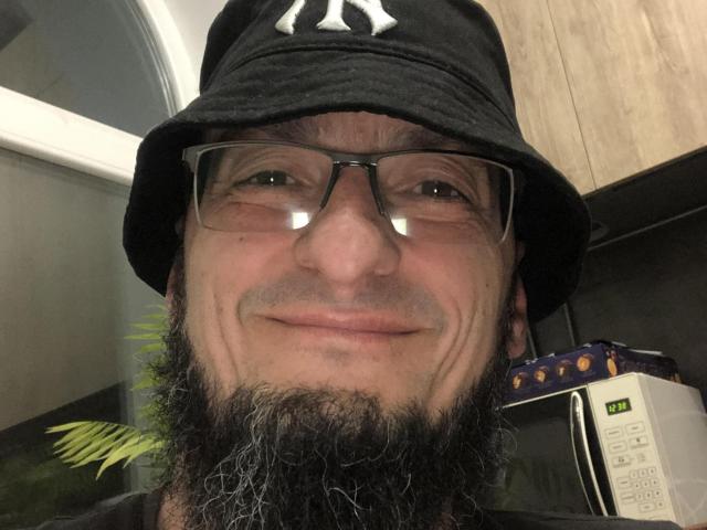 BernardFunny with a black bucket hat, glasses, and a hairy beard, smiling at the camera. His nose is slightly upturned and he has a relaxed expression.