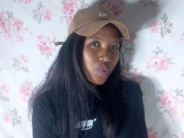 KattyMiriam with dark skin, long black hair, wearing a brown cap and black shirt, pouting slightly, looking at camera. Her tits are small and she has a cute face.