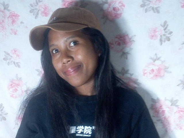KattyMiriam with dark skin, long black hair, brown cap, black shirt, smiling at camera, showing small ass, cute face, pink floral wallpaper in background.