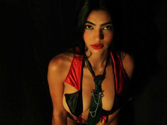 Joy Firestone, avec ses cheveux foncés, son rouge à lèvres rouge et sa cravate verte, portant un haut rouge et noir découpé qui expose ses seins, sur un fond noir, regardant directement l'appareil photo..