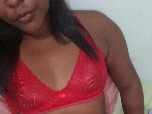 SabrinaBrown69 with dark skin wearing a red lace bra, showing her small tits, a tiny red bow in the center, and a subtle smile, with black hair.