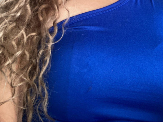DalilaSuperHot's blue, shiny, tight-fitting top highlights her big, round, and slightly sweaty tits. Her curly blonde hair is on the left, partially in the frame.