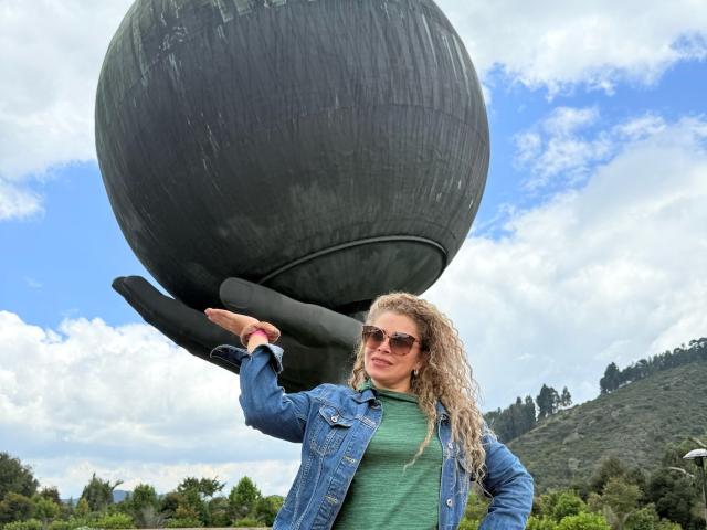 Dalila, com cabelos loiros cacheados, usando óculos de sol, uma camisa verde e um casaco jeans azul, levantando uma grande escultura de bola preta com base preta. O céu está azul com nuvens brancas..