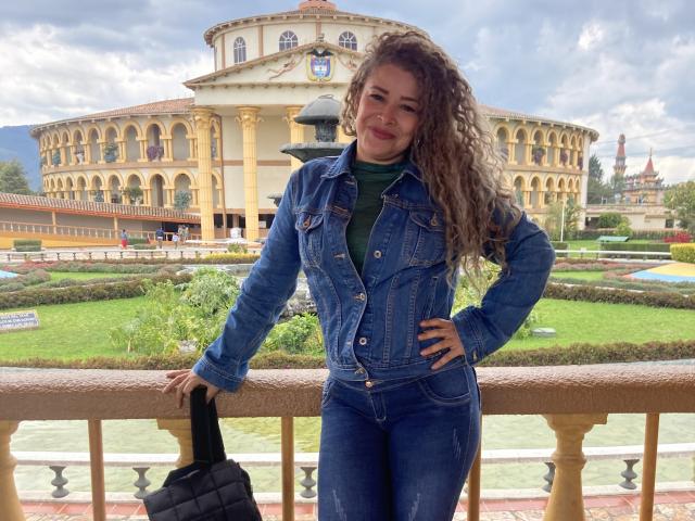 Dalila, con el pelo castaño rizado, vistiendo una chaqueta vaquera azul y una camisa verde, de pie con una mano en la cadera, sosteniendo un bolso negro, sonriendo, frente a un edificio amarillo..