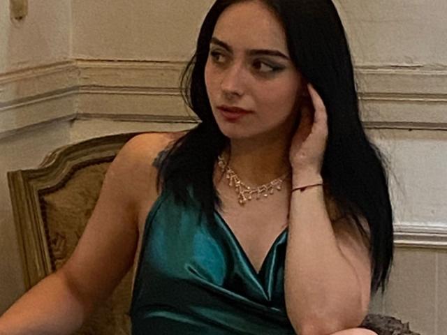 AshleyDaphna, com longos cabelos pretos, vestindo um vestido de cetim verde brilhante, um colar e uma pulseira vermelha, olhando para o lado com um leve sorriso, com a mão tocando sua orelha..
