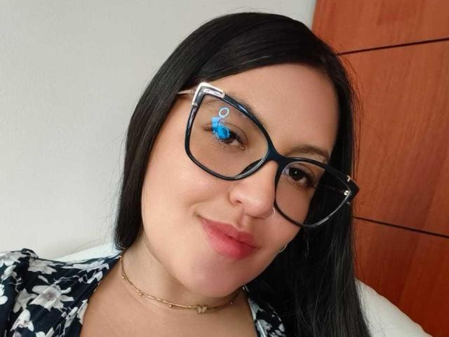 NathDawnson has black hair, blue eyes with a small blue dot on her right eye, wearing big black glasses, a floral shirt, and a gold chain, smiling softly.