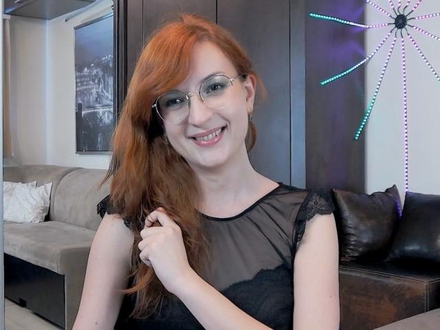 MoonBlossom with red hair, glasses, and a black sheer top, holding her hair, smiling at the camera, showing her white teeth, and a cute nose. Boobs visible under top.