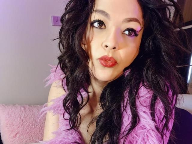 KarenCurlyHair with long, wavy black hair, wearing a pink feathered top, has red lipstick and thick eyelashes, pouting her full, glossy pink lips, looking slightly to the side.