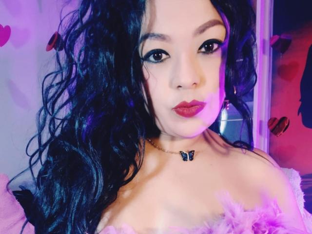 KarenCurlyHair has long black curly hair, red lipstick, and a butterfly necklace, wearing pink fluffy off-shoulder top, with purple and pink lighting on her face.