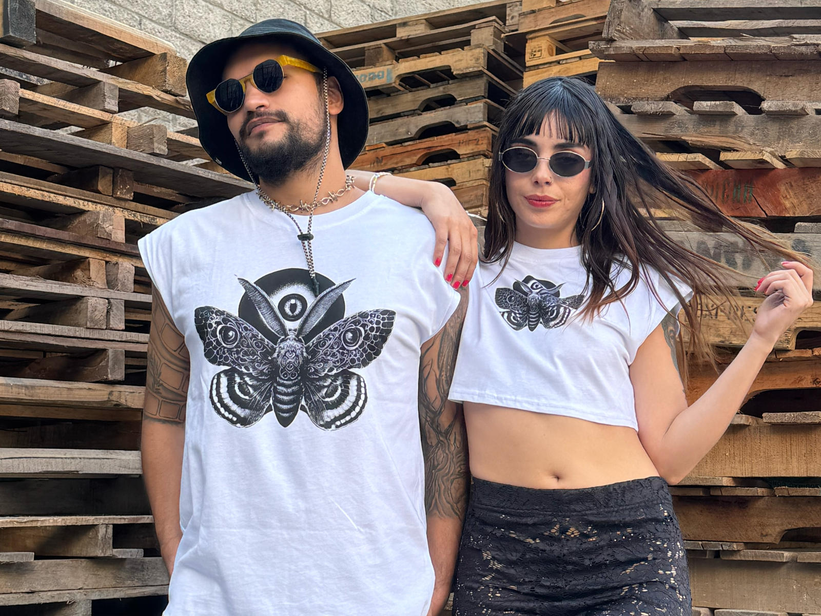 ErhosAndPsyche (man) with beard, black hat, sunglasses, white tee, butterfly graphic, and ErhosAndPsyche (woman) with black lace pants, white tee, butterfly graphic, and hand in hair.