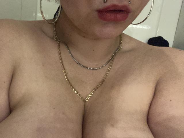PixiiieKitty ha la pelle chiara, rossetto rosso, un piercing al naso, grandi orecchini a cerchio dorati e una collana a doppia catena, che mostra i suoi seni scoperti e una collana d'oro..