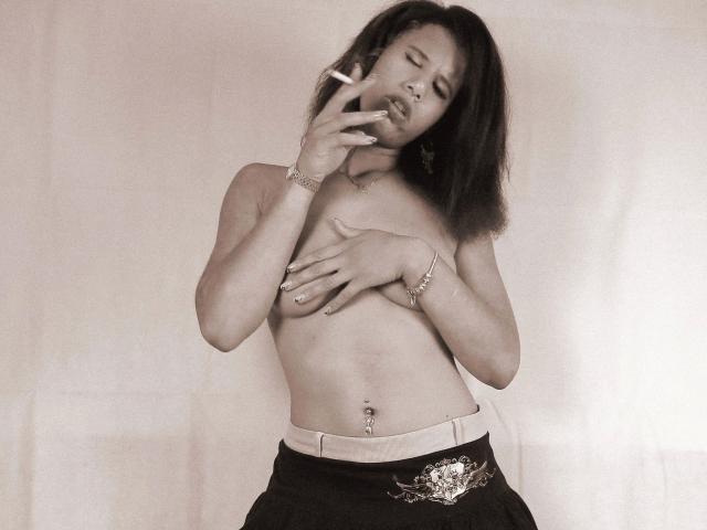 AaliyahHarris-hot is topless with small tits, covering her right boob with her hand, wearing dark pants and a belt with a floral design, holding a cigarette to her mouth.