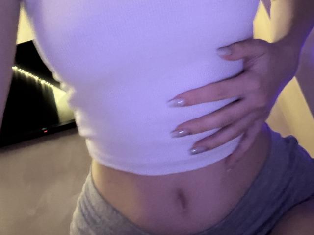 A camiseta branca de SelinaRose, que mostra sua barriga plana e a mão com unhas longas e pintadas em tons de nude, usando shorts cinzas, em um quarto com pouca luz..