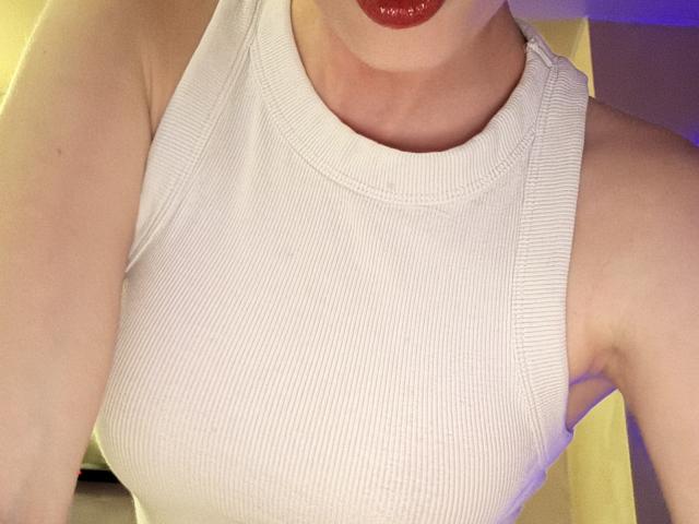 La camiseta blanca con textura de SelinaRose, que muestra sus pechos, con lápiz labial rojo en el labio inferior, y sus brazos ligeramente levantados, en una habitación iluminada con luz amarilla..