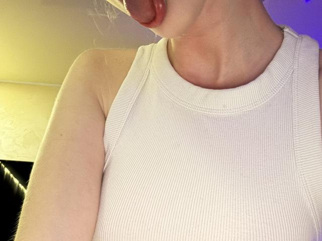 SelinaRose, con la lengua afuera, vistiendo una camiseta blanca de tirantes con textura, mostrando su hombro descubierto y parte superior del pecho. Su lengua está ligeramente extendida, tocando su labio inferior..