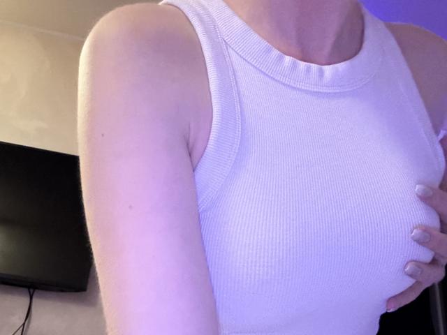 A blusa regata branca texturizada da SelinaRose, que mostra seus ombros e peito nus, com a mão dela segurando suavemente seus seios. Suas unhas estão pintadas em uma cor clara..