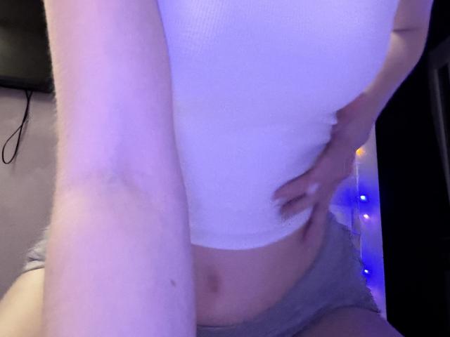 SelinaRoses weißes, geripptes Tanktop, das ihren Bauch freilegt, und graue Shorts, wobei ihre Hand auf ihrem Unterbauch liegt und eine leichte Bräune sowie eine violettliche Hautfärbung sichtbar sind..