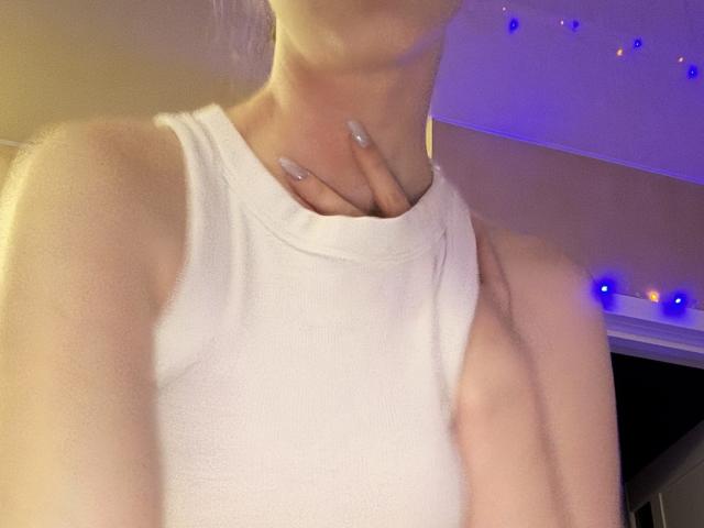 SelinaRose vestindo uma blusa branca sem mangas, sua mão tocando suavemente o pescoço, exibindo suas unhas prateadas bem cuidadas, sob uma iluminação roxa suave..