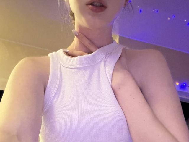 SelinaRose, de piel pálida, vestida con una camiseta blanca de tirantes, con la mano en el cuello, luces LED azules y moradas en el fondo, labios ligeramente entreabiertos, su rostro parcialmente visible..