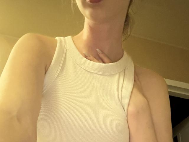 SelinaRose, com a pele clara, vestindo uma camiseta branca de tirantes, mostrando o peito e o pescoço, com a mão perto da garganta, e os lábios levemente entreabertos..