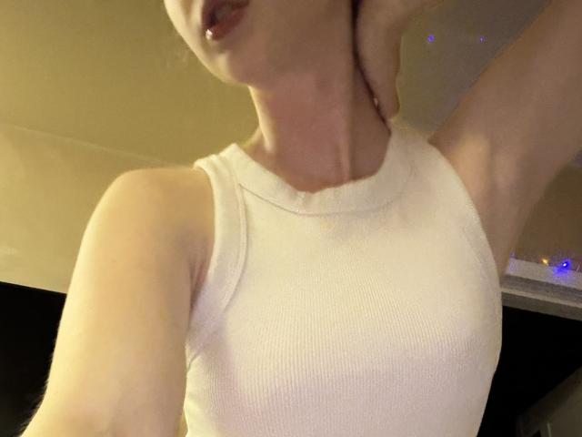 SelinaRose portant un débardeur blanc côtelé, avec une lèvre percée visible, un bras levé et une peau claire, montrant ses petites seins et ses fesses légèrement visibles..