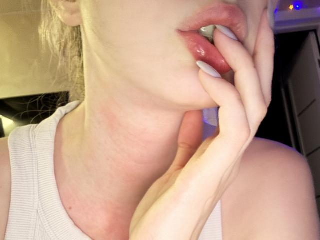 SelinaRose's face with her hand near her glossy pink lips, white nails, and a pale neck, wearing a white tank top, in a slightly angled selfie.
