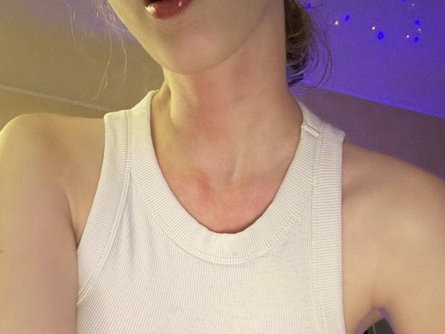 SelinaRose, com a pele clara, lábios carnudos, vestindo uma camiseta branca de tirantes, mostrando a clavícula e a parte superior do peito, com sua pele rosada visível no pescoço..