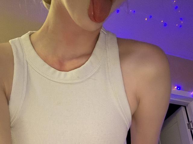 SelinaRoses weißes Rippstrick-Tanktop, mit herausgesteckter Zunge, zeigt ihre nackten Schultern und ihren Schlüsselbein. Ihr Gesicht ist teilweise außerhalb des Bildes. Sie hat helle Haut..