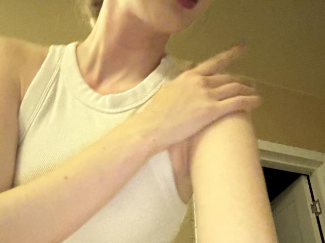SelinaRose with fair skin, wearing a white sleeveless top, touching her upper arm with her right hand, visible from neck to elbow. Her face is partially shown.