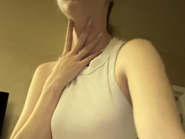 SelinaRose with her hand on her neck, wearing a white ribbed tank top, showing her tits and a bit of her ass, in a dimly lit room.