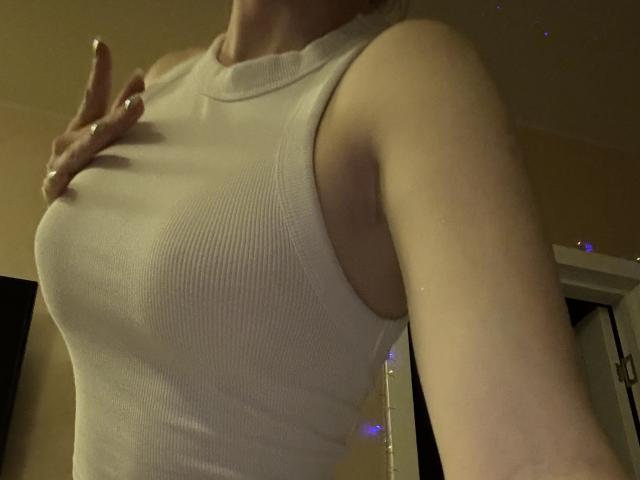 SelinaRoses weißes, geripptes Tanktop zeigt ihre kleinen, festen, wohlgeformten Brüste. Sie macht mit ihrer rechten Hand ein Friedenszeichen, und ihr Arm ist leicht angehoben..