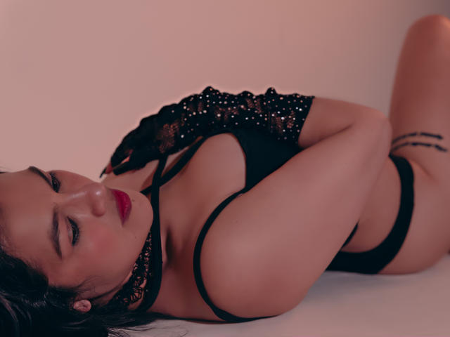 ElizabethDupont lying on her side, wearing black lace gloves and lingerie, with her tits and ass exposed, her red lips slightly parted, and a tattoo on her right thigh.