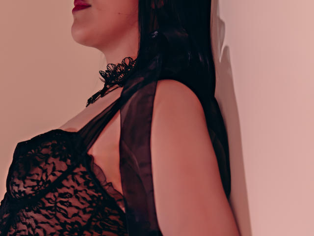 ElizabethDupont with black lace lingerie, showing her tits in a sheer top, choker, and red lipstick, leaning against a wall, her face partially visible, dark hair.
