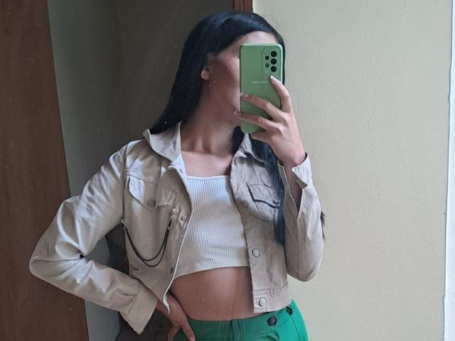 HathorX with long black hair, wearing a beige jacket over a white ribbed crop top, green pants, holding a green phone, covering her face, showing her slim, tan midriff.