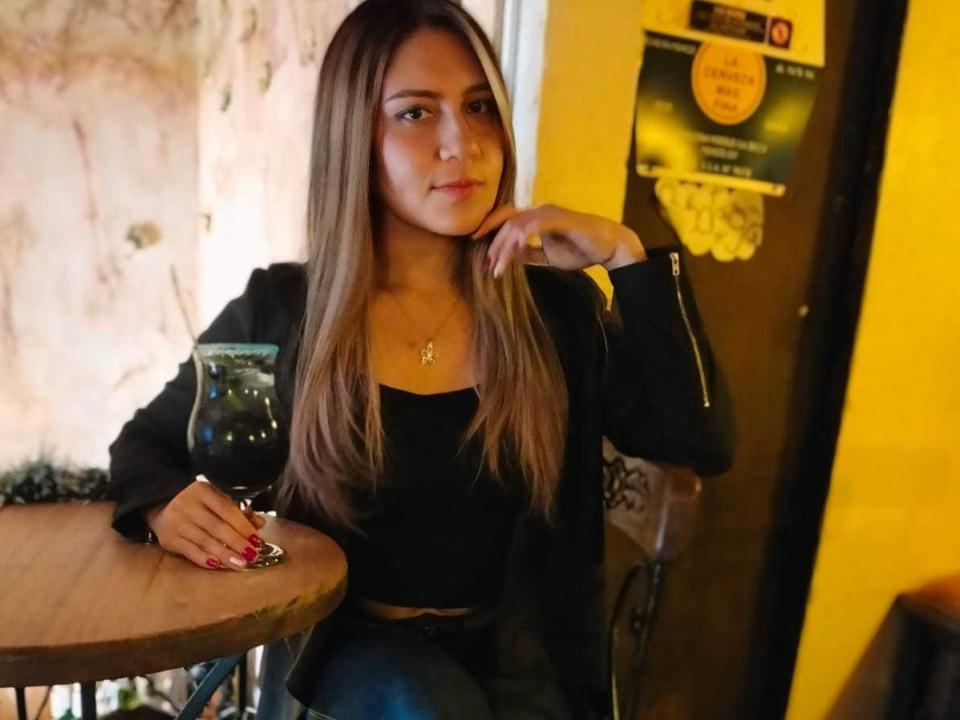 CinthiaRoss with long brown hair, black top, black jacket, and blue jeans, holding a glass of dark liquid, sitting at a wooden table, yellow wall in background.