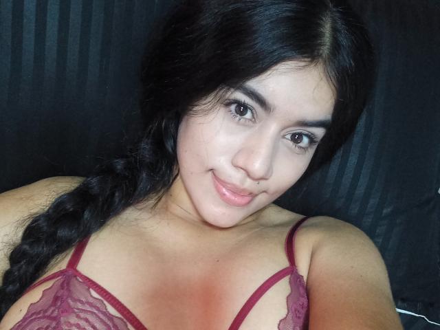 IsabellaTasty with dark hair in a braid, wearing a red lace bra, smiling at the camera, showing her tits. Her skin is light, and she has a cute face.