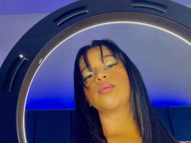 AntonelaTawer has dark hair, green eyeshadow, and pink lips, looking up with a slight pout, reflected in a circular mirror with a blue light behind.