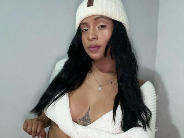 AntonelaTawer with long black hair, wearing a white knit beanie and off-shoulder white top, showing her tits and a tattoo under her chest, with a neutral expression.