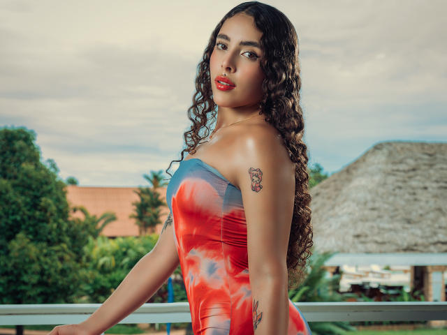 TianaVera with long curly brown hair, red lipstick, and a tight strapless dress with red, blue, and pink swirls. She has tattoos on her arm and shoulder, looking sexy.