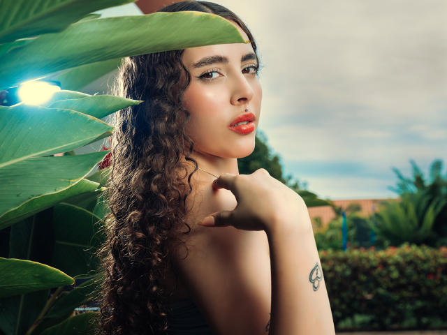 TianaVera with curly brown hair, red lipstick, and a tattoo on her right forearm, pointing to her chin, standing in front of green leaves with a bright light.