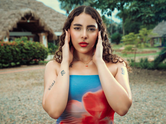 TianaVera with curly brown hair, wearing a blue and red strapless dress, hands on her face, tattoos on arms, red lipstick, standing outdoors, gravel ground, tropical background.