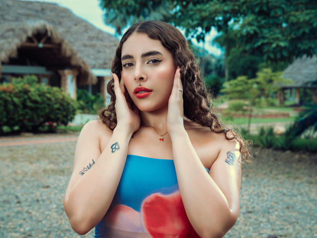 TianaVera has curly brown hair, red lipstick, and blue strapless dress. She's touching her face, showing tattoos on her hands and arms. Her boobs are visible. Outdoor setting.